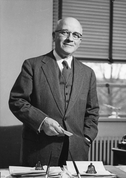 J. Harold Lampe wearing glasses and dressed in a three piece suit standing behind his desk.