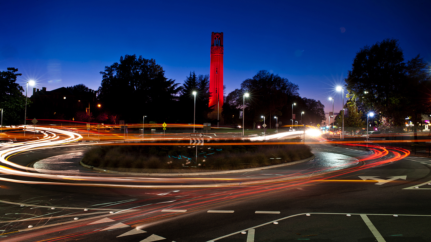 Time-lapse image of NC State belltower in red light at dusk with automobile headlight lines navigating the adjacent traffic circle.