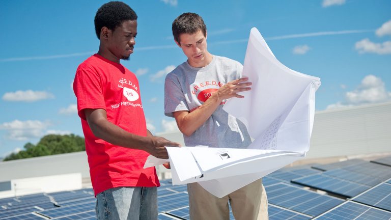 Student photovoltaics researchers stand over a bank of solar panels, studying plans.