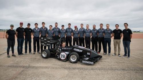 Group of 17 standing behind small F1 style racing car.