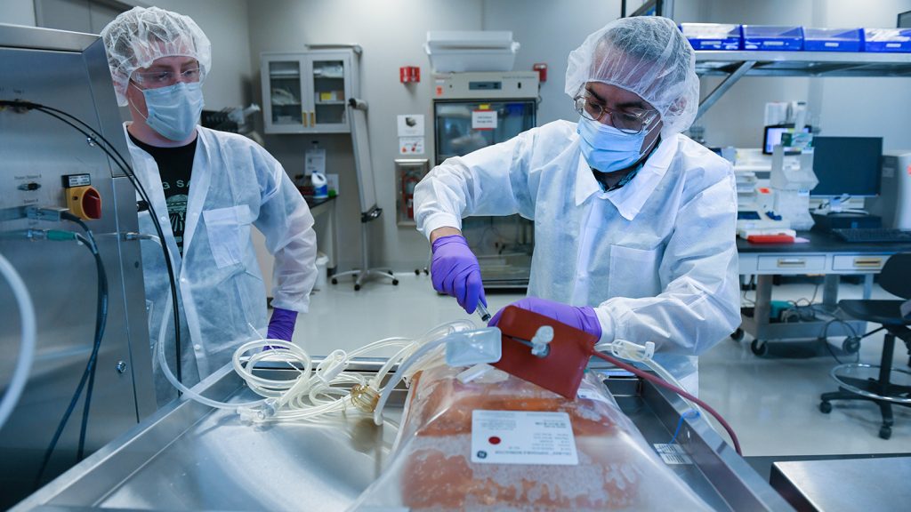 Two workers dressed in full lab gear working at an industrial table.