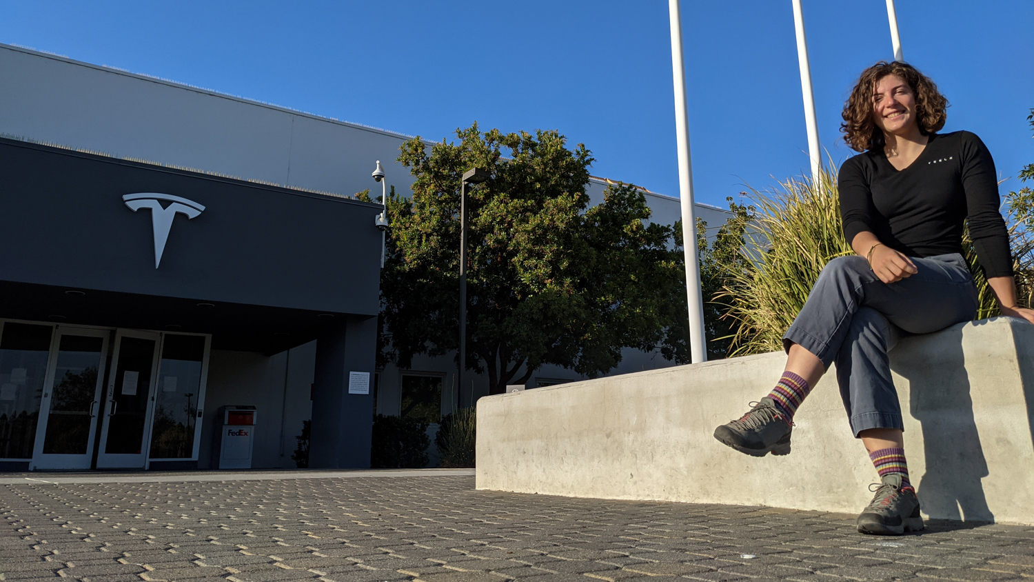 Morgan Mase sitting on concrete wall in front of building adorned with Tesla logo.