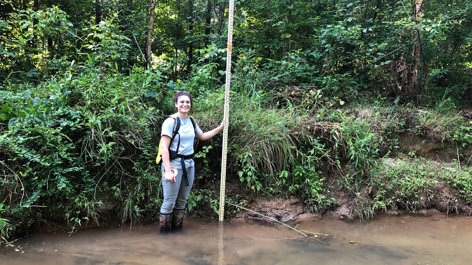 Layla El-Khoury in waders standing in stream next to bank holding a tall measuring stick.