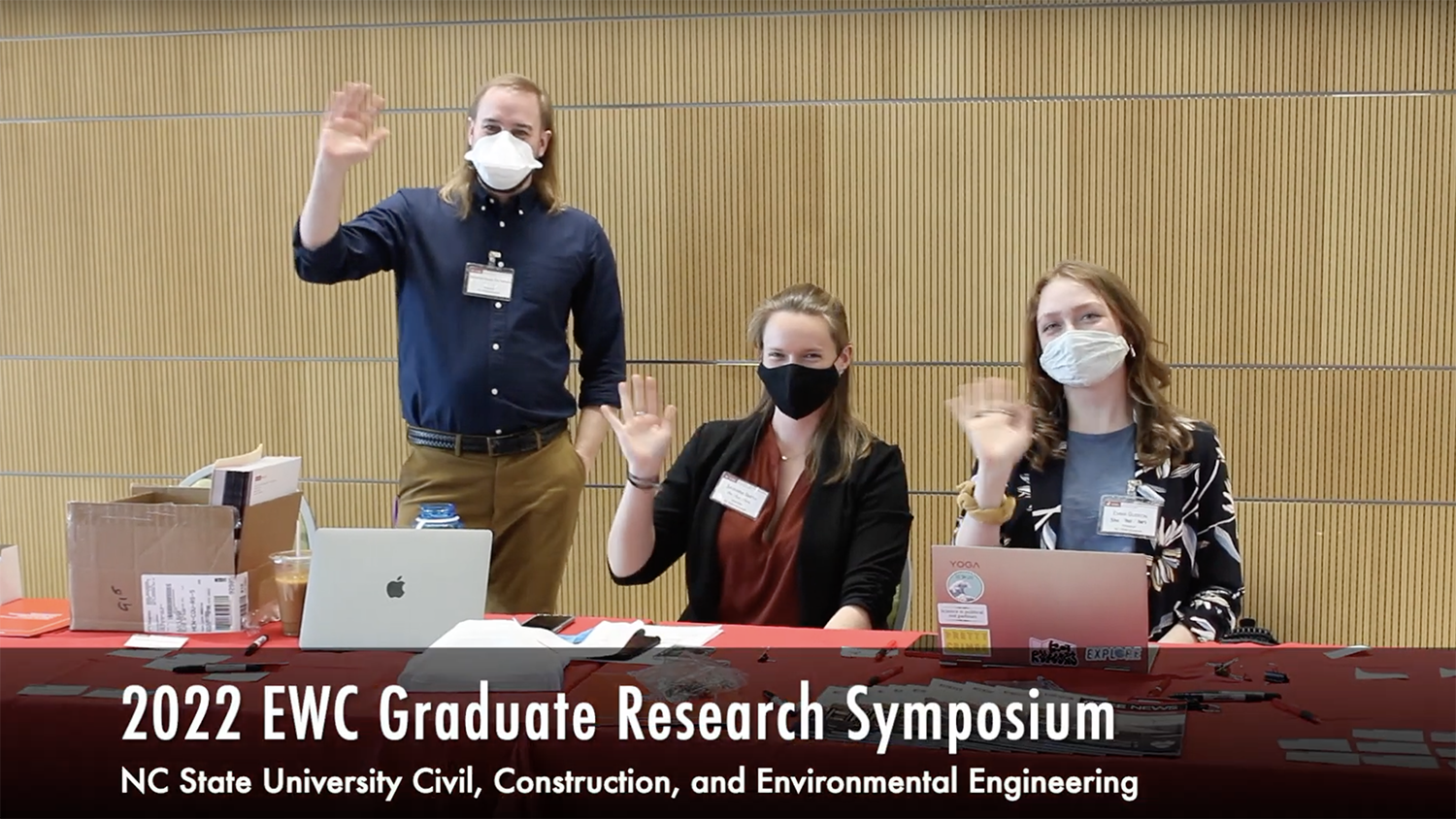 Photo of graduate students waving at camera from behind a presentation table.