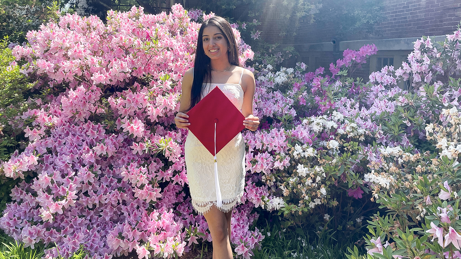Manali Shirsekar, wearing a white dress while holding a red graduation cap, poses in front of pink azaleas.
