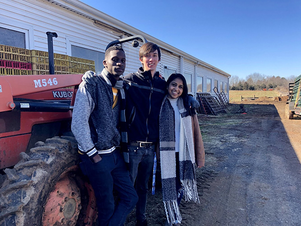 From left, Ziwa Mukungu, Matthew Simpson, Shraddha Rathod. All three are standing in front of a red tractor and a long white building.
