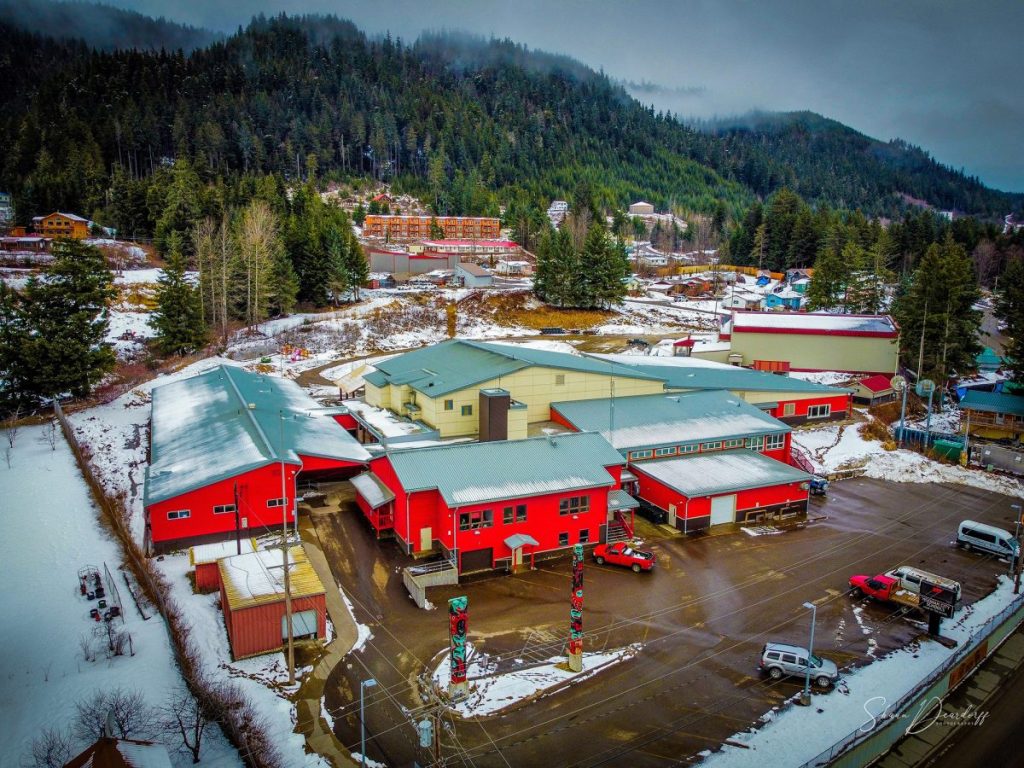 Aerial view of Hoonah School, red buildings with green roofs, thin layer of melting snow on ground with mountain ridge and evergreen trees in background.