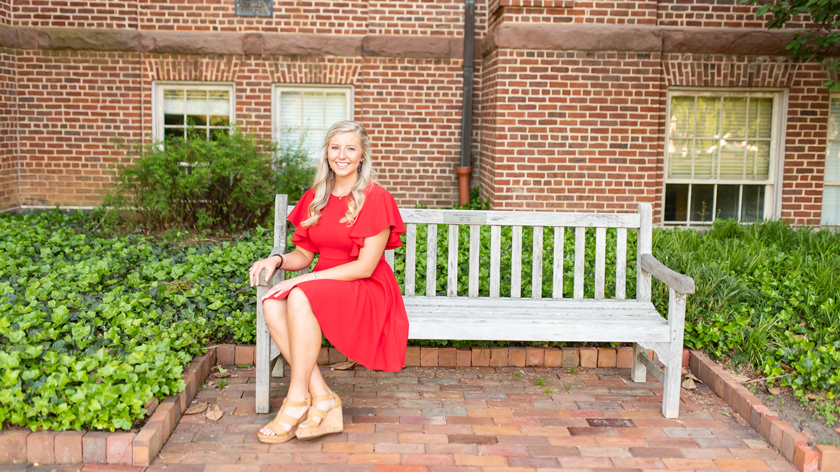 Emory New, wearing a red dress, poses for picture while sitting on a wooden bench.