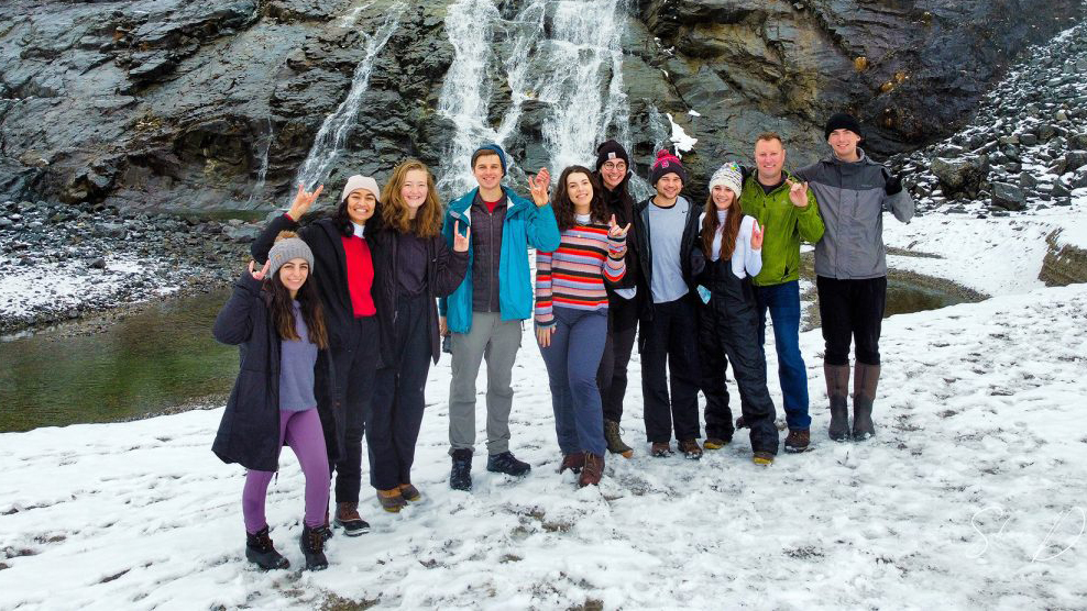 Group of NC State students pose together in front of Alaskan waterfall.