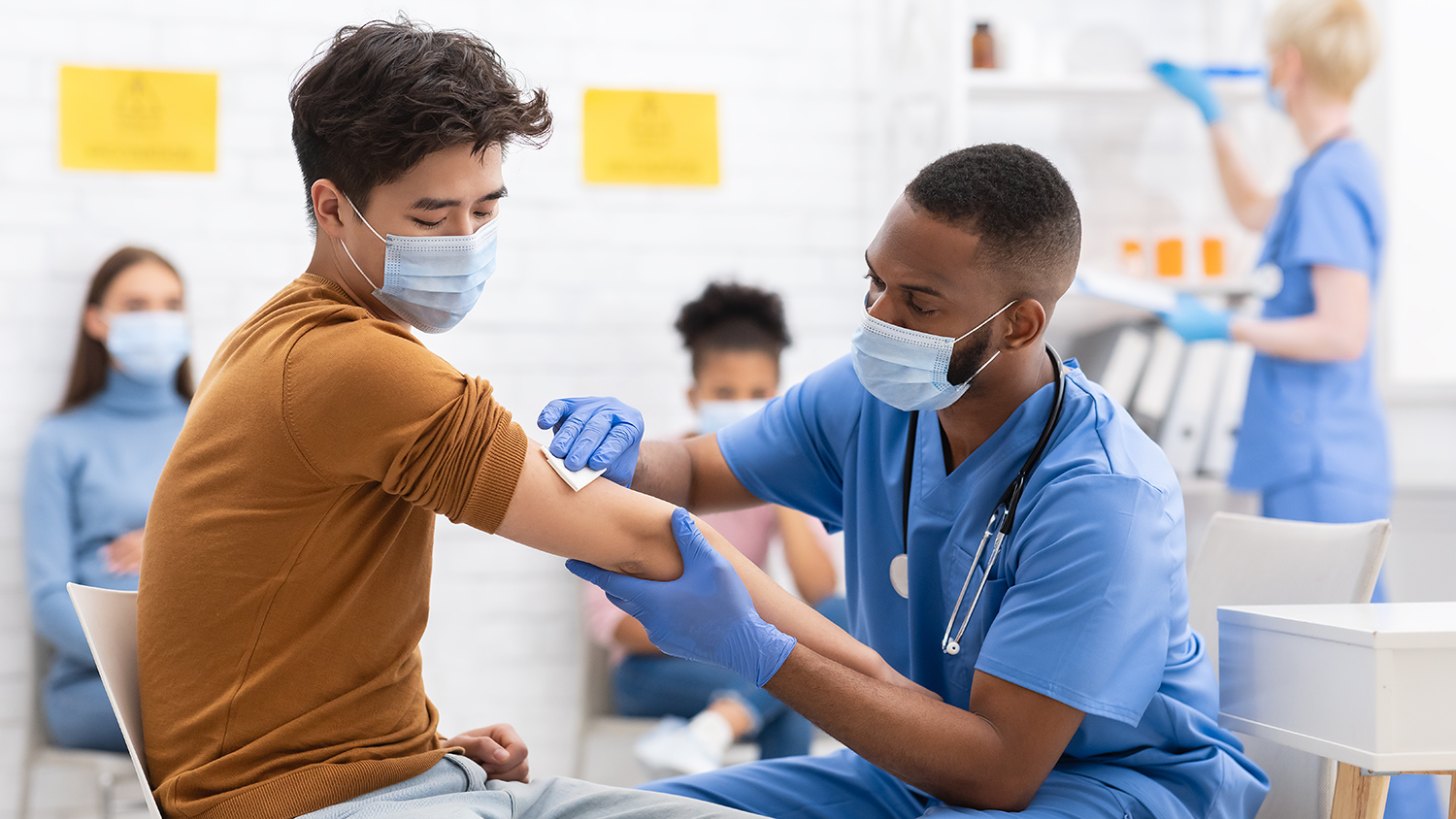 Healthcare professional dressed in blue scrubs administers vaccination to seated patient.