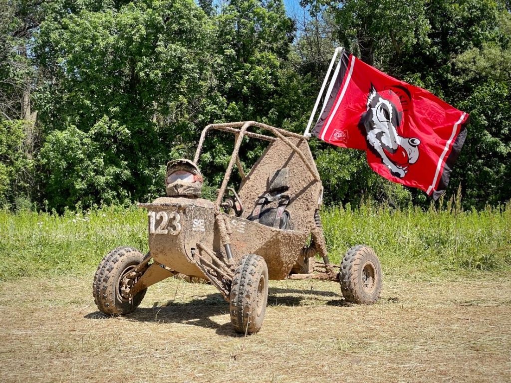 NC State University Pack Motorsports Baja SAE Team vehicle covered in mud and displaying the NC State wolf flag in red.