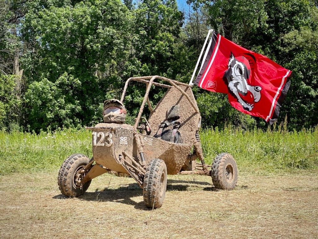 NC State University Pack Motorsports Baja SAE Team vehicle covered in mud and displaying the NC State wolf flag in red.