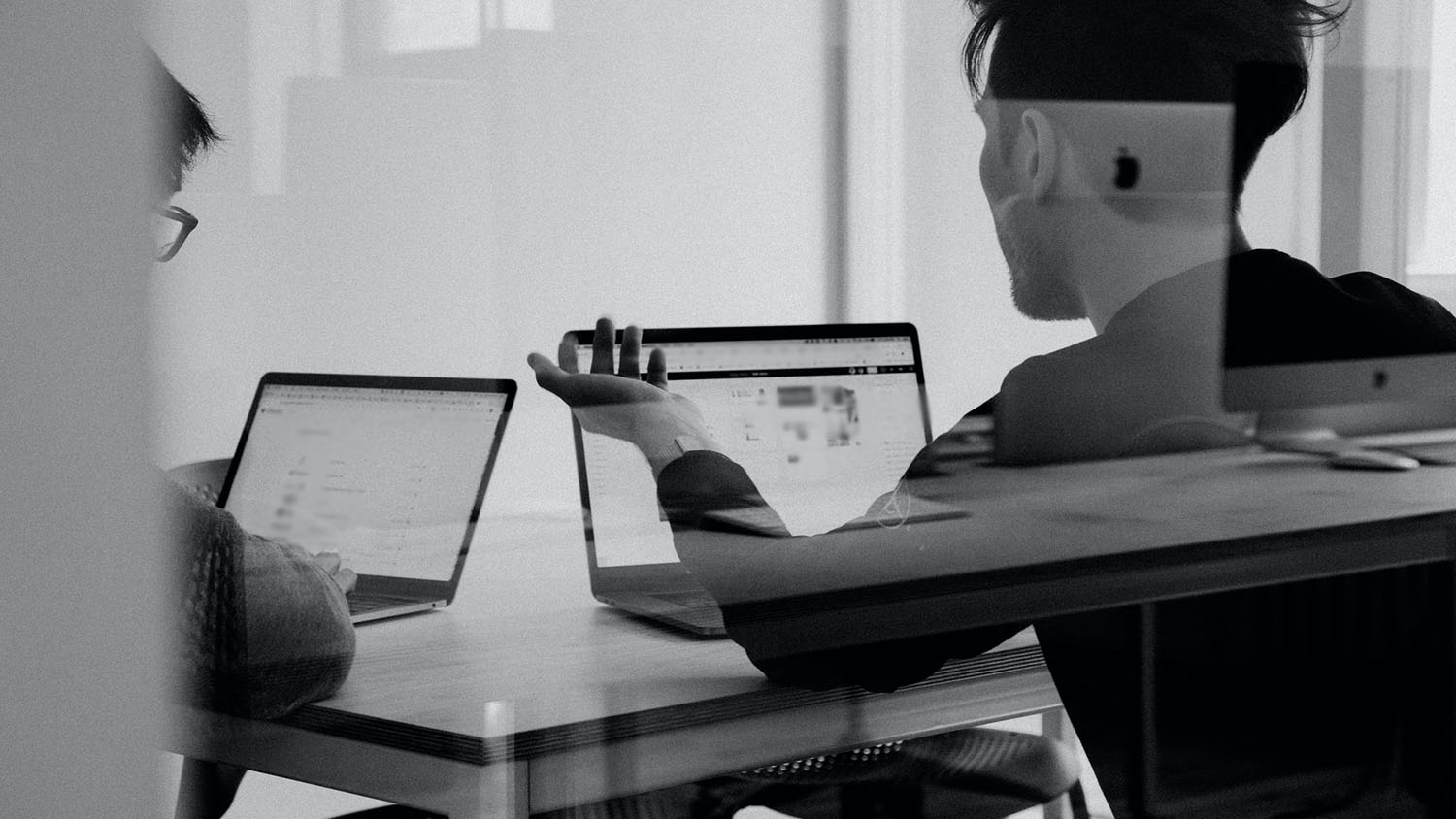 Black and white photo of back of two people engaged in conversation while seated in front of laptops.