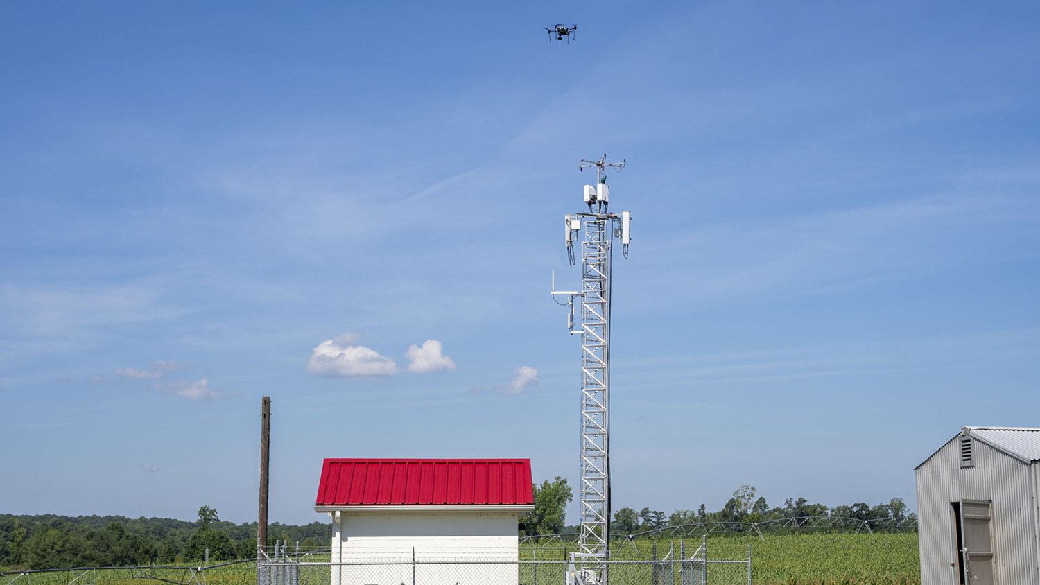 Small black drone hovers over field and small white building with red roof.