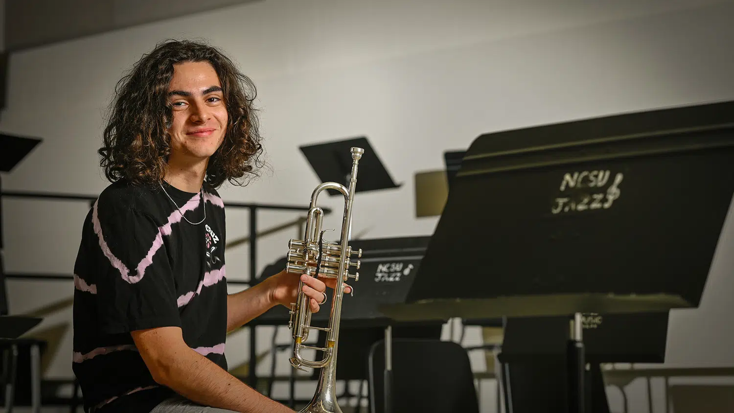 Aidsan Dumain looks at camera while holding a trumpet and seated in front of a music stand.