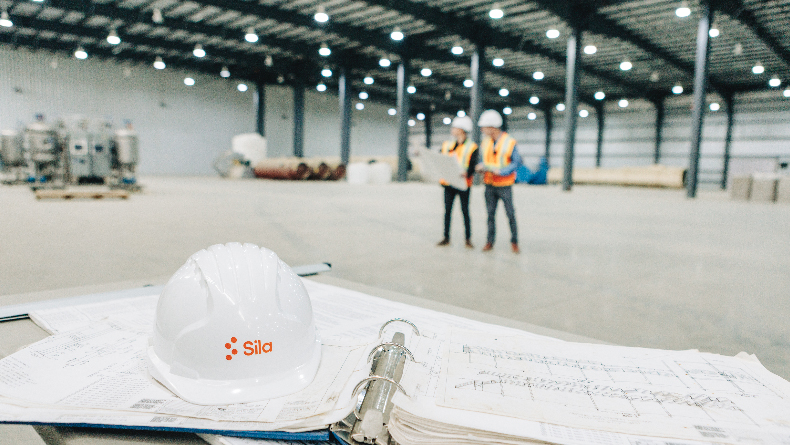 Image of Sila white hardhat on table with papers in the foreground and two employees in the background.