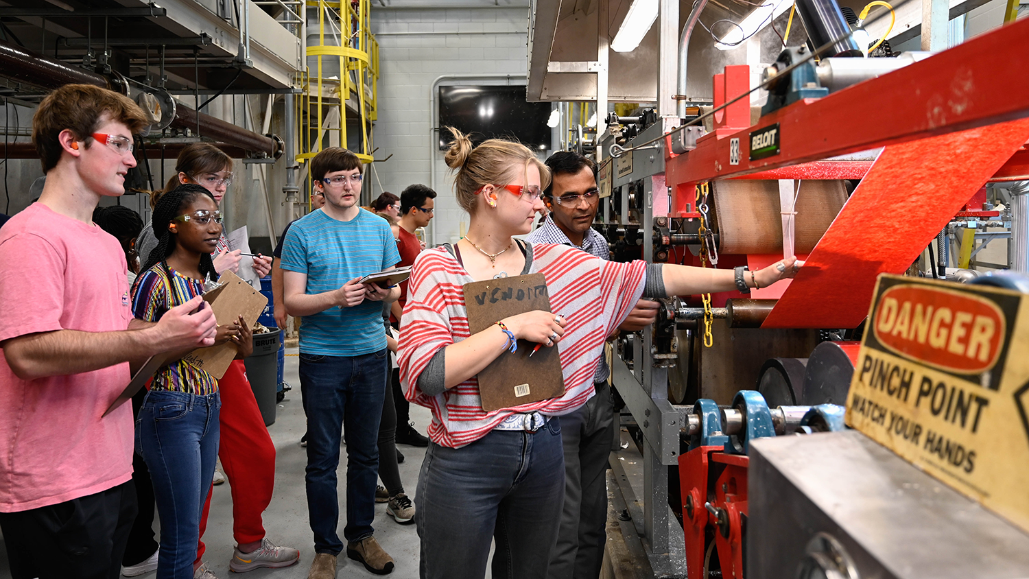 A group of students each wearing clear safety glasses listen and watch as a professor shows one of the students the manufacturing line for a roll of red paper.