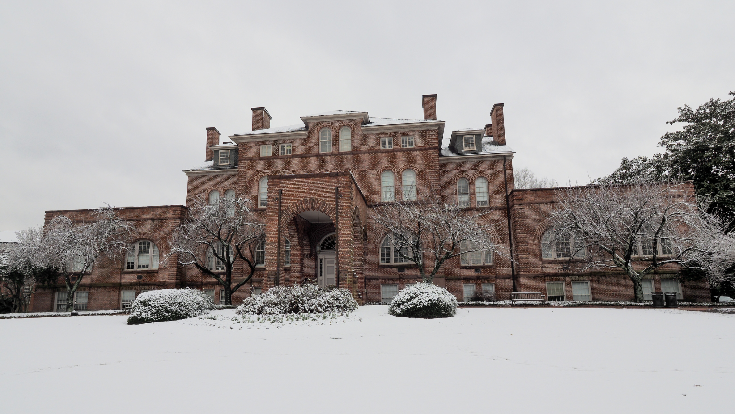 Holladay Hall, an all brick colonial-style building, with a blanket of white snow on the ground, trees and shrubbery and a light grey, overcast sky.