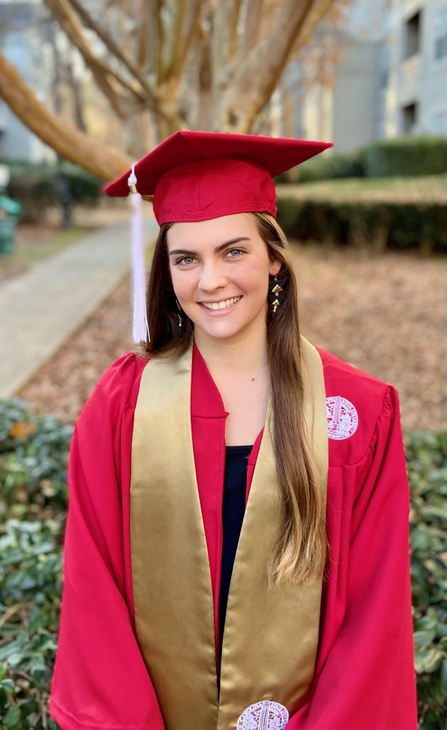 Lynne Dale dressed in red graduation cap and gown with fall trees and leaves in the background.