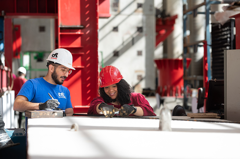 Male and female students work in the Constructed Facilities lab on Centennial Campus. Male on left is wearing a white hardhat and a blue tshirt. Female on right is wearing a red hardhat and red shirt.
