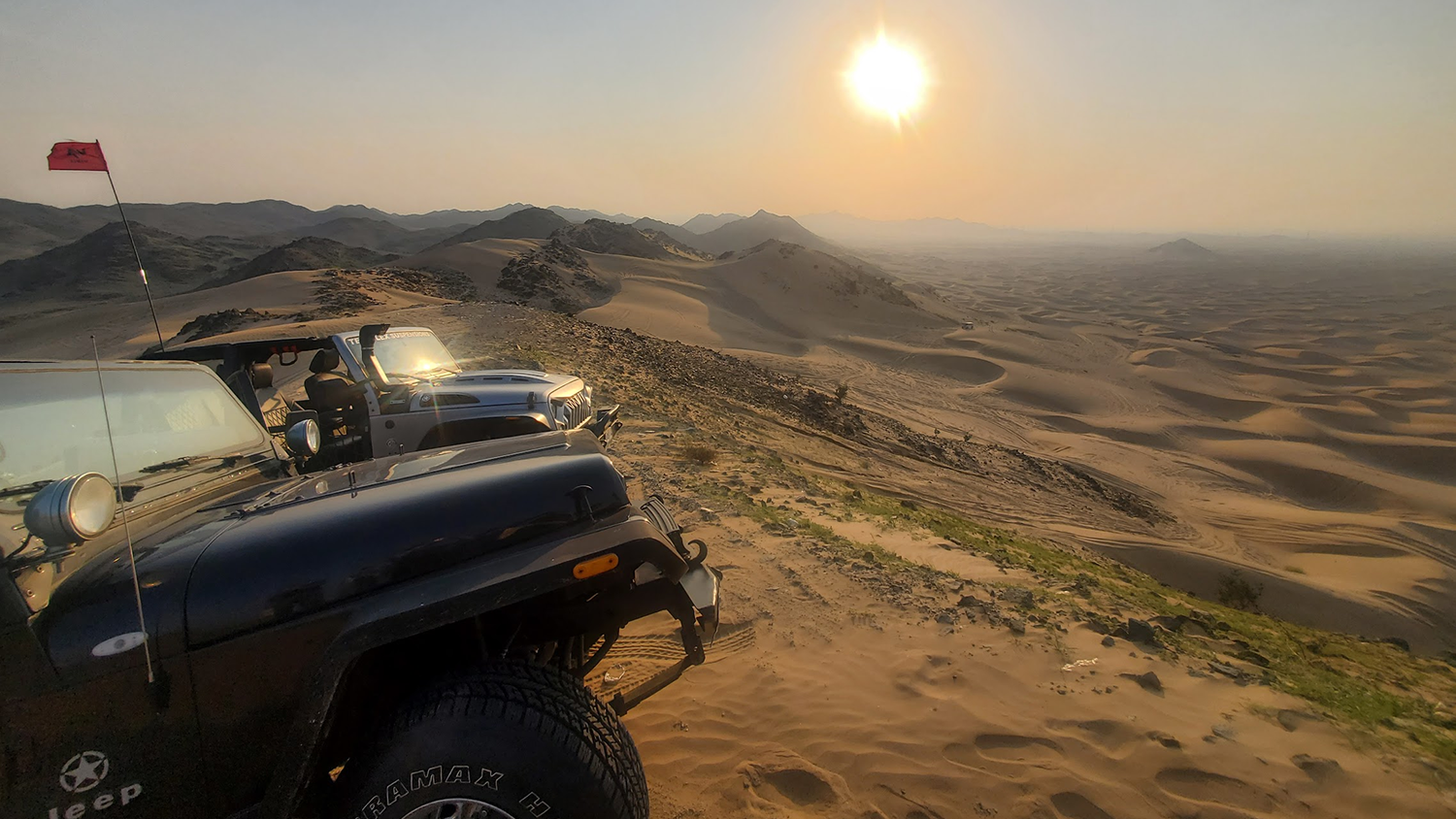 Two Jeeps, left, sit atop a sand dune ridge facing the sunset in the distance.