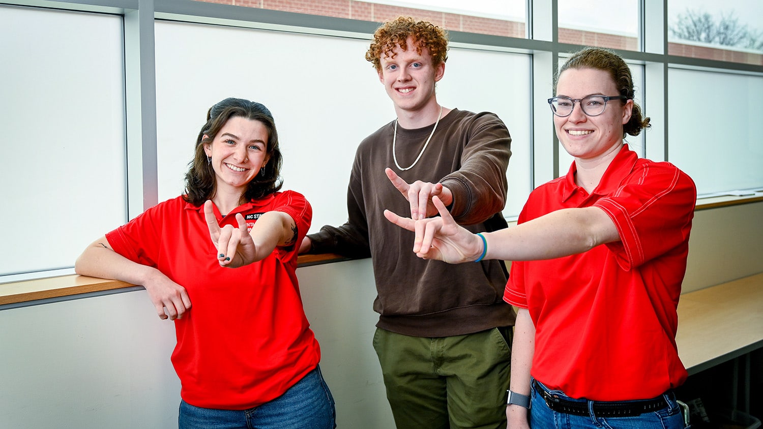Alexis Jacobs (left, wearing red short-sleeve shirt), Caleb Moore (middle, wearing dark olive green long-sleeve shirt) and Lauren Bradley (right, wearing red short-sleeve shirt) are three of NC State's Mental Health Ambassadors. All three are showing the Wolfpack hand sign.