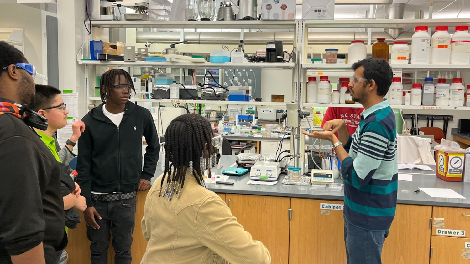 High school students listen to a professor while gathered in a laboratory.