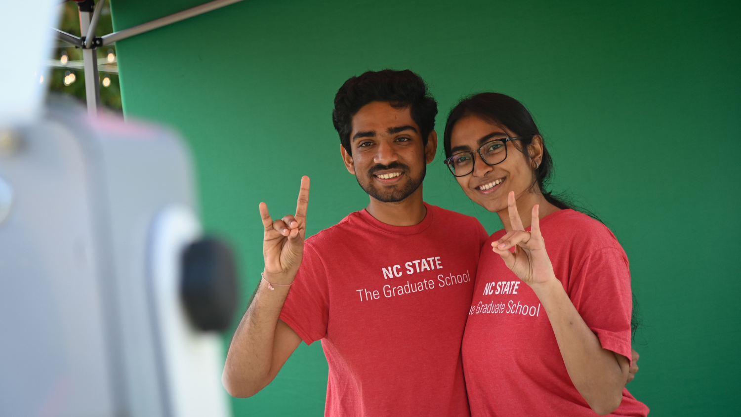Two students dressed in red t-shirts stand in front of a green screen and flash the Wolfpack hand sign.