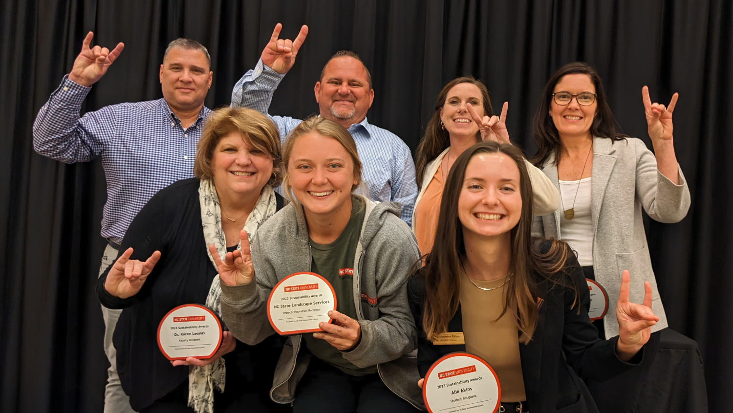 Winners of NC State's Sustainability Awards pose for a group picture while flashing the Wolf hand sign. They are standing in front of a black fabric background.