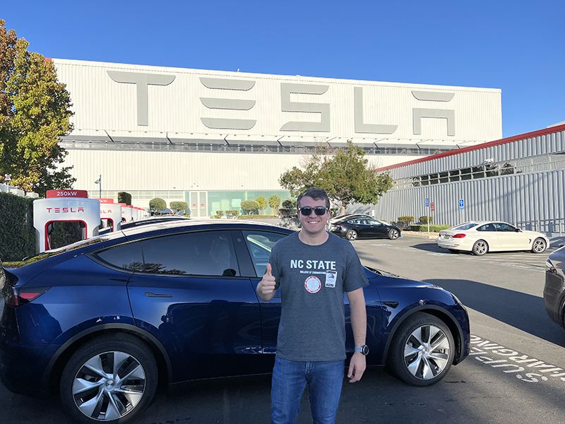 Robert Brenneman poses in front of a Tesla vehicle and manufacturing facility.