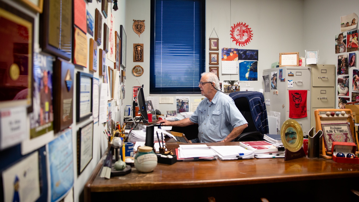 Al Tonelli sets in front of his desk in his office and works at his computer. His office walls are covered in awards, diplomas and photos.