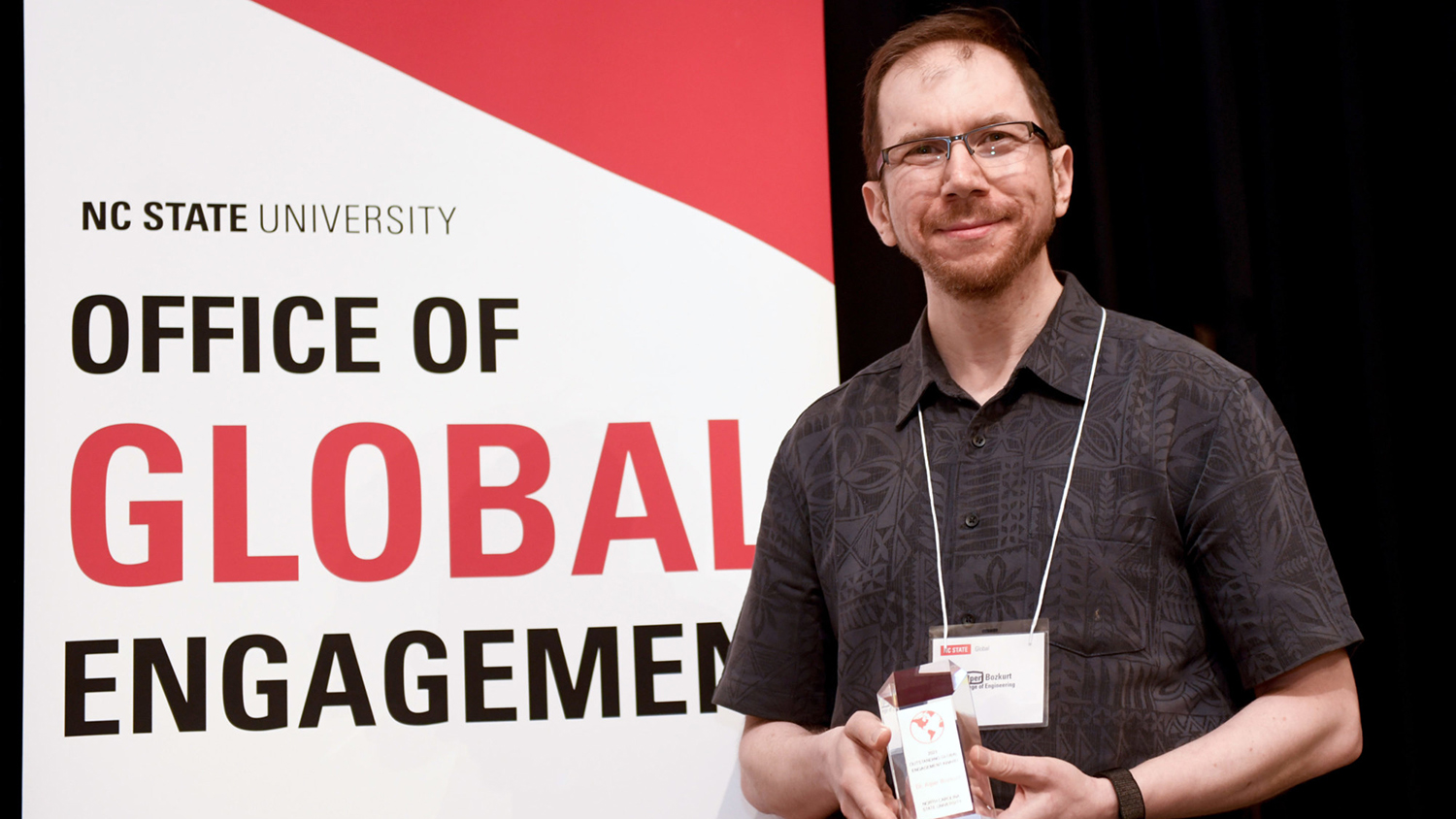 Alper Bozkurt holds his award whiile posing for a photo in front of the Office of Global Engagement banner.