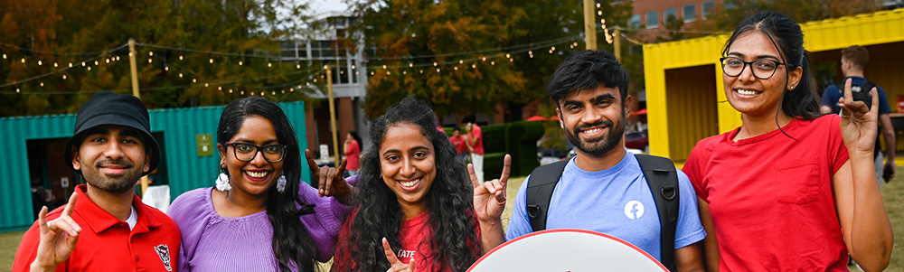 Group of 5 students pose for picture while displaying the NC State wolfpack hand sign.