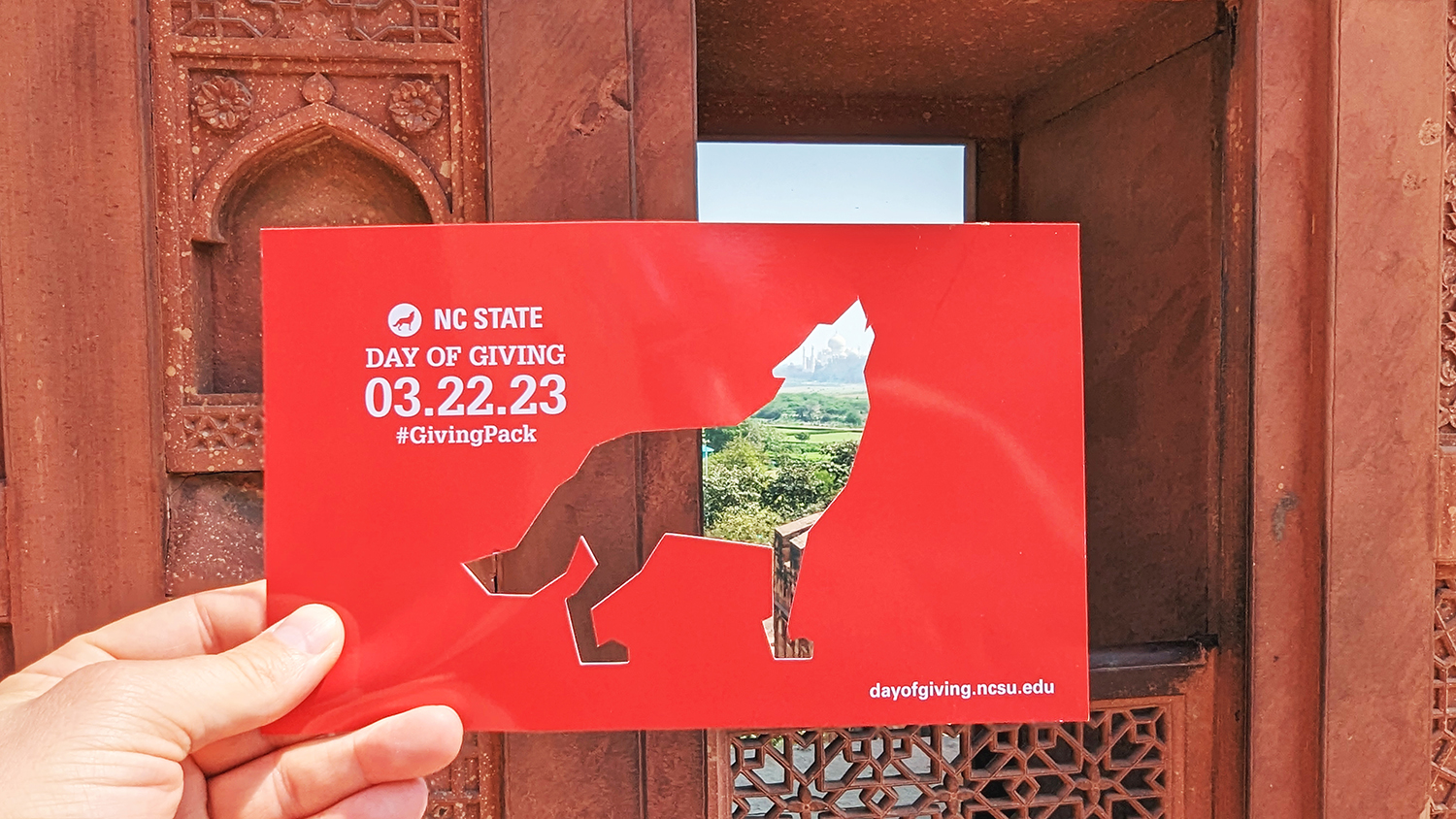 Hand holding a red card with an outline of a howling wolf cutout so as to see the view of the brick building and landscape behind. Day of Giving 3/22/2023 #GivingPack is written in white letters on the card.