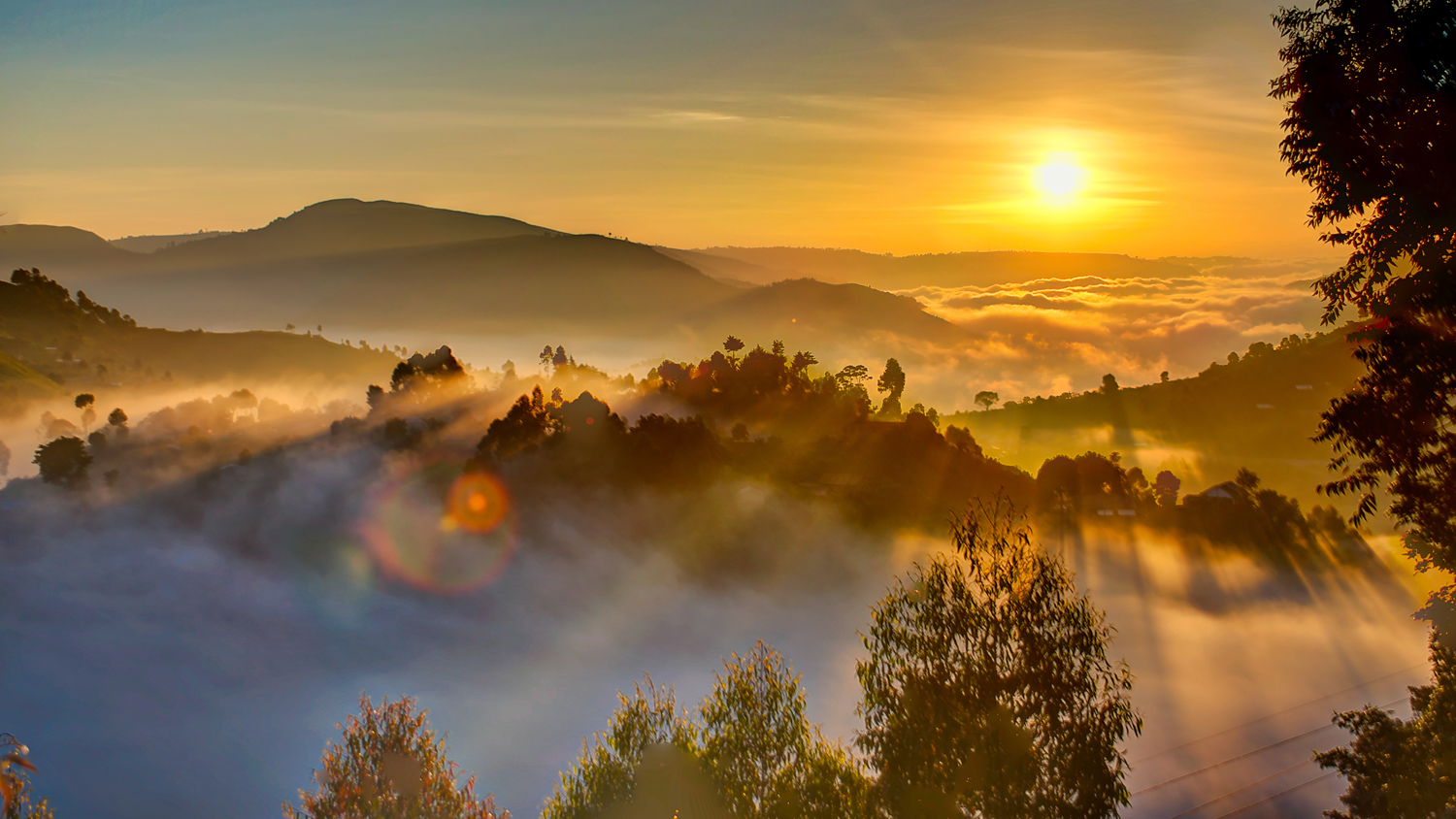 A late evening aerial view of the Kampala, Uganda landscape. A small mountain peak surrounded by low clouds wrapped around the treetops is on the left and the setting sun is on the right.