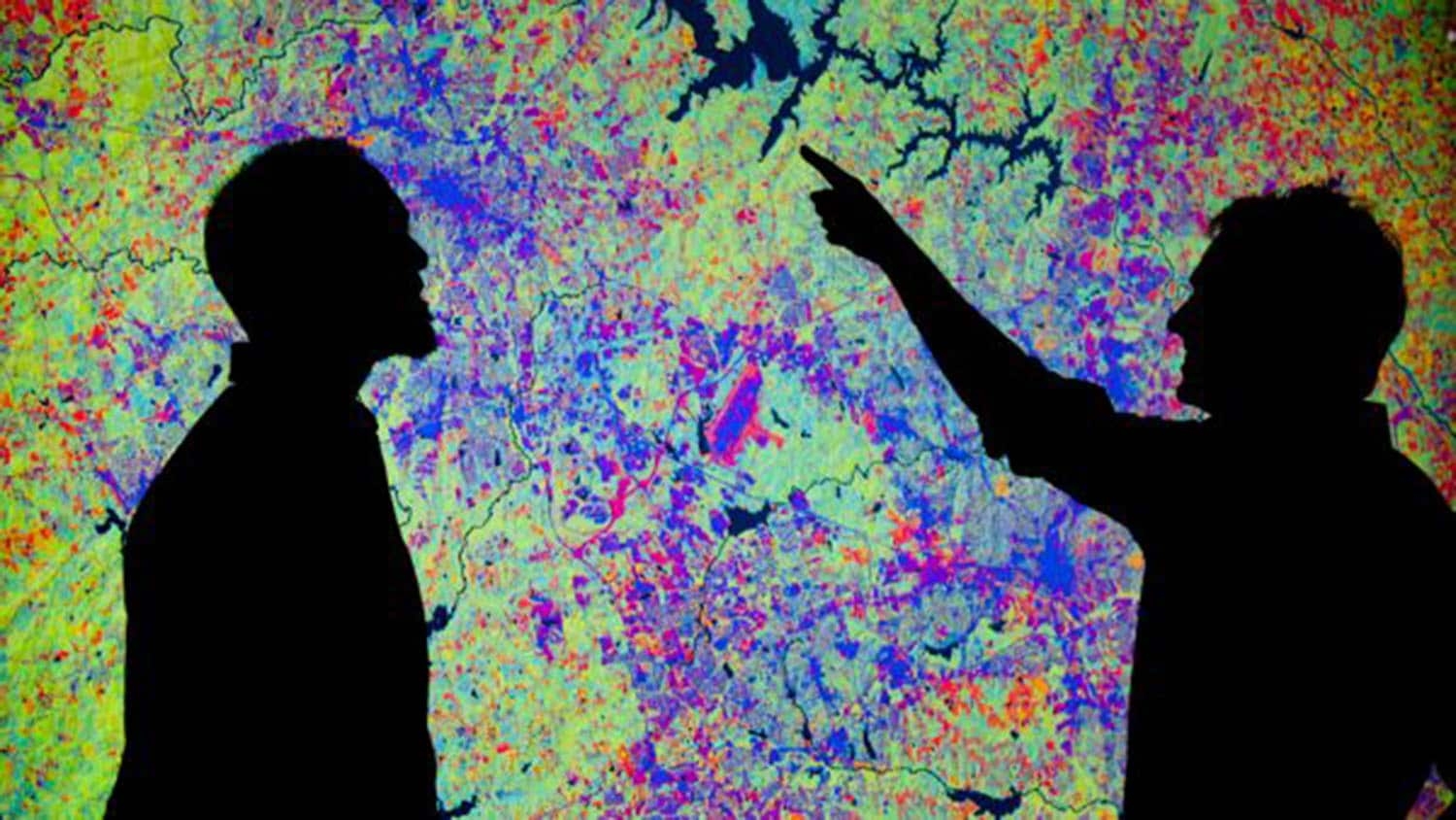 Two researchers appear as silhouettes in front of a screen showing a digital heat map filled with greens, blues, reds, oranges and various other shades of colors.