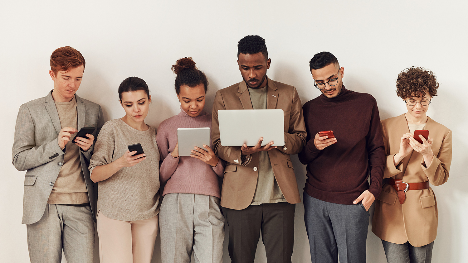 Group of people leaning against a wall using a variety of digital devices.
