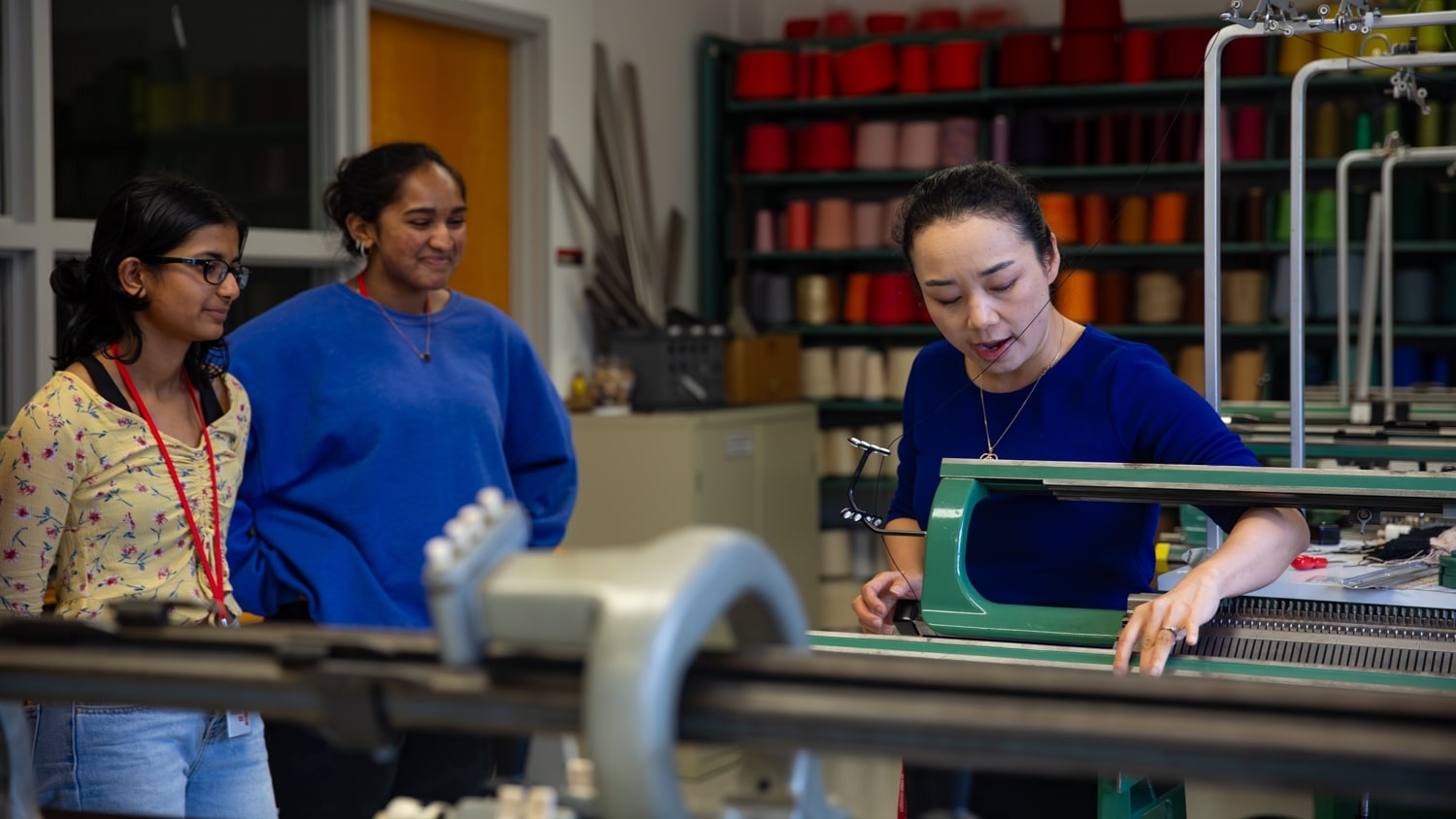 Xiaomeng Fang stands behind a knitting machine and points to a certain part of the machine to show two students to her left how it works.