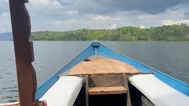 Photo taken from inside of front of an open-air boat painted blue with green tree-lined shoreline in the distance.