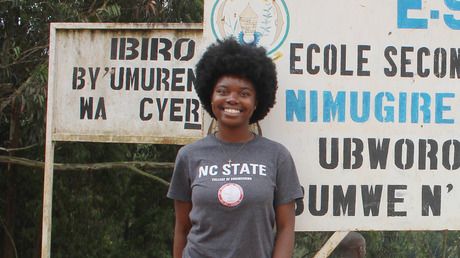 Jenni Mangala wearing a grey NC State t-shirt stands in front of a Rwandan secondary school.