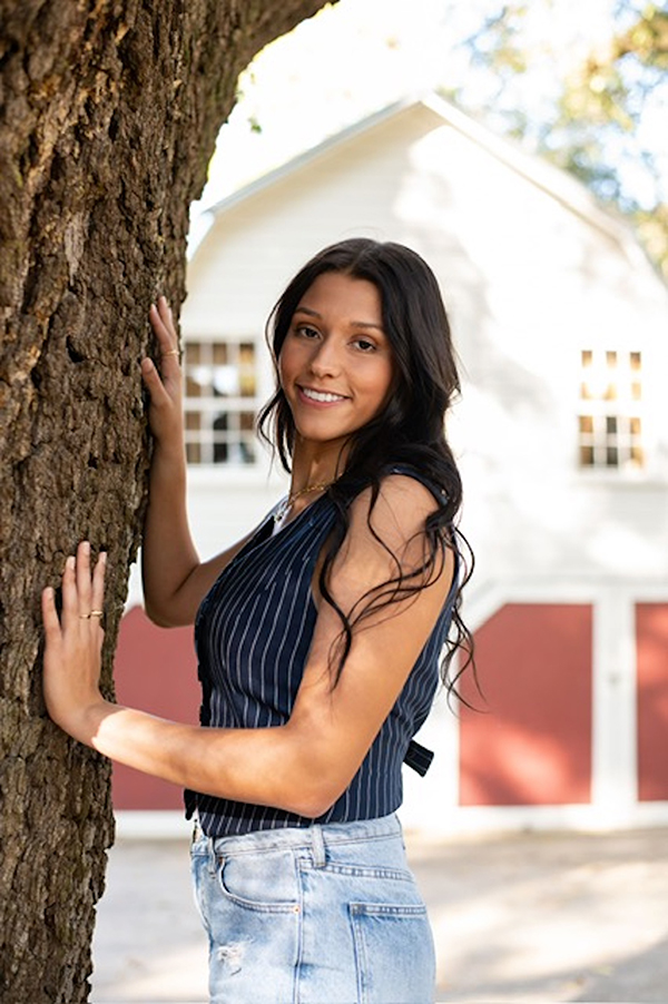 Raven Cummings poses leaning on tree. A white building with red doors can be seen in background.