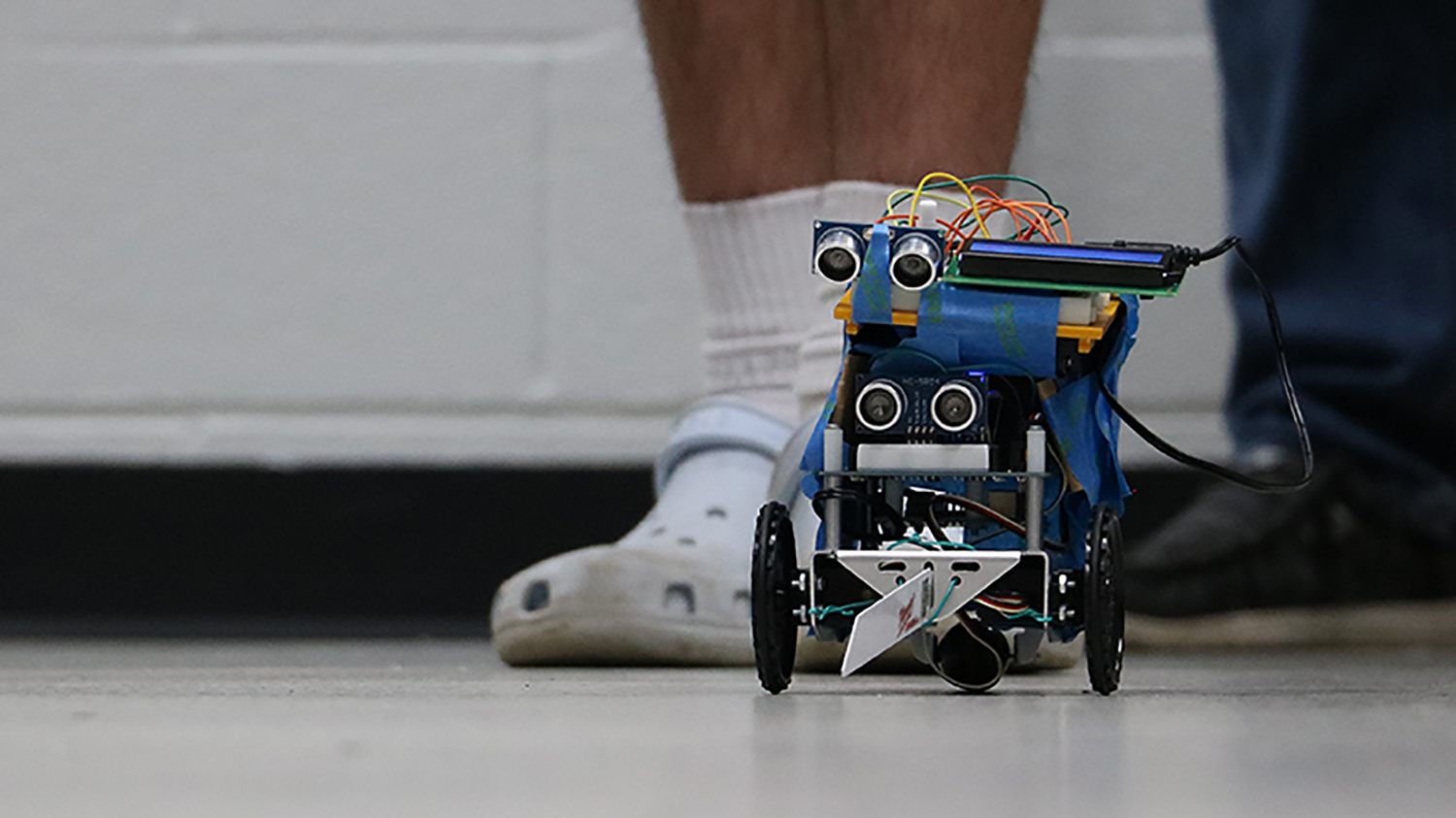A floor level photo of a small blue-colored , black-wheeled agricultural robot in use at a summer camp. The floor is white and there are two sets of shoes in the background behind the robot.