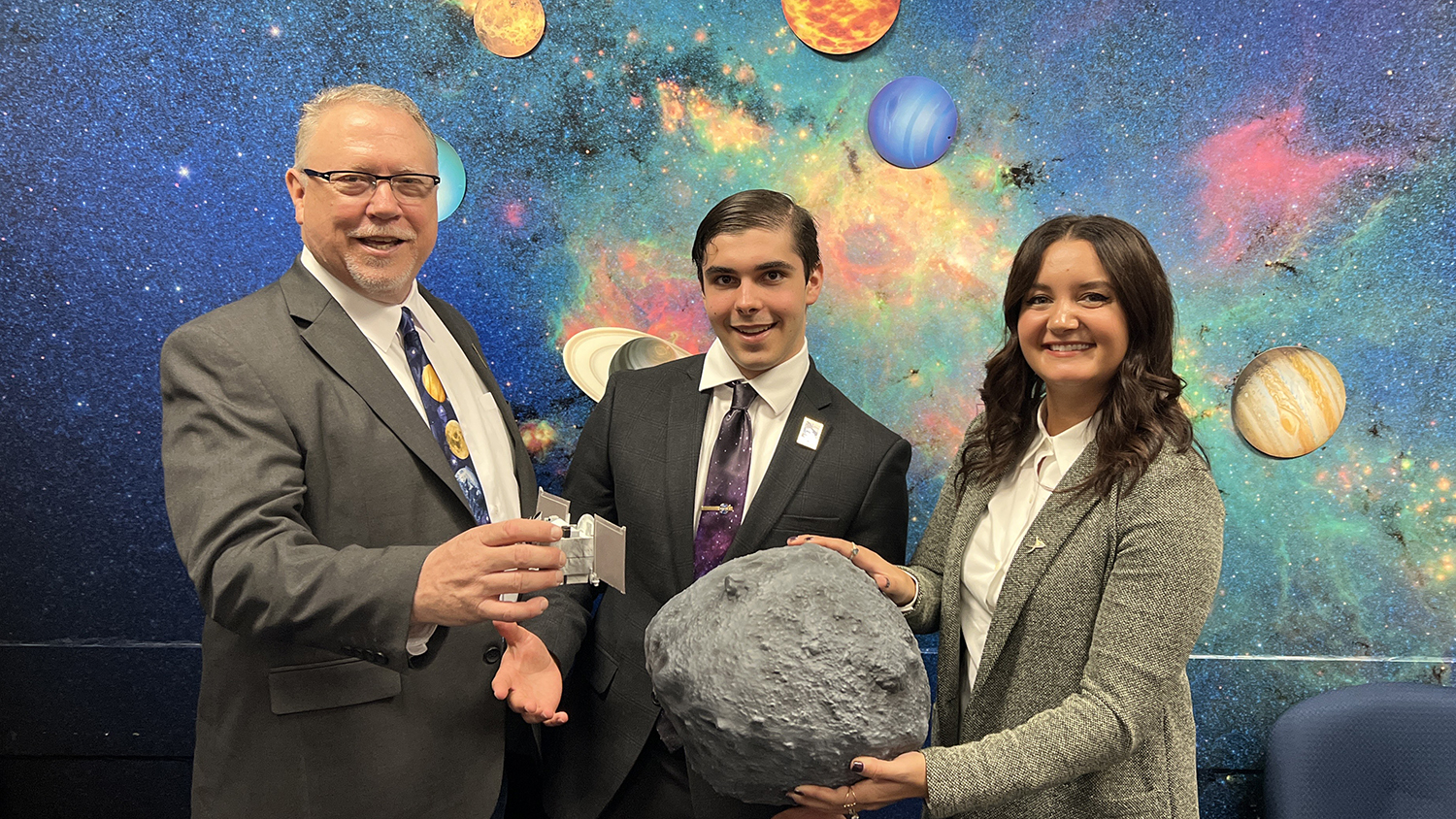 Michael Puzio, center, helps hold a piece of asteroid. On his left is a man and on his right is a woman, both helping to hold the asteroid fragment. Behind all three is a colorful mural depicting various celestial bodies and objects.