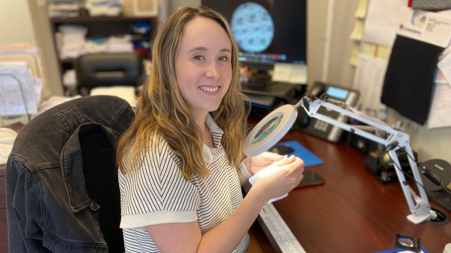 Hannah Orlowski sitting at her desk holding a swatch of fabric under a microscope.