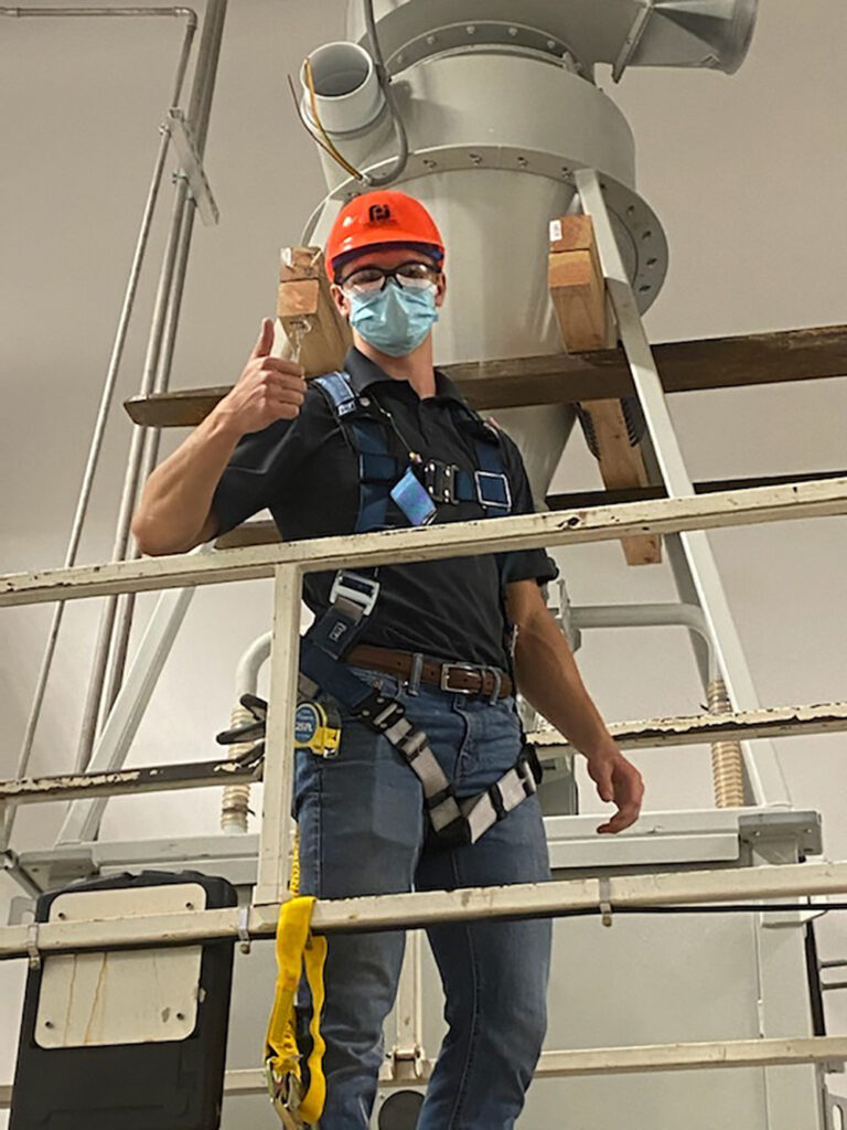 Caleb Jolley, wearing a red hardhat and face mask, gives the thumbs up from an overhead walkway in an industrial space.