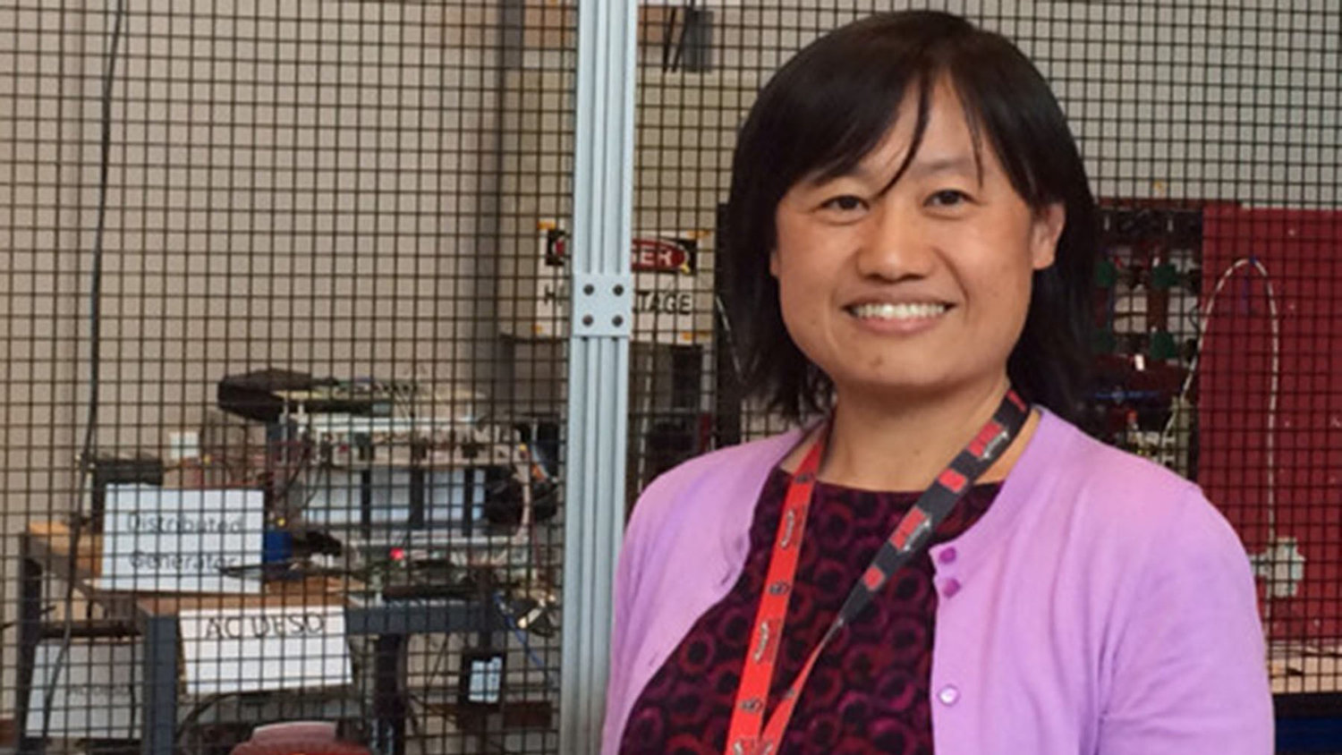 Ning Lu, wearing a lavender cardigan sweater and red lanyard, stands in front of laboratory equipment.