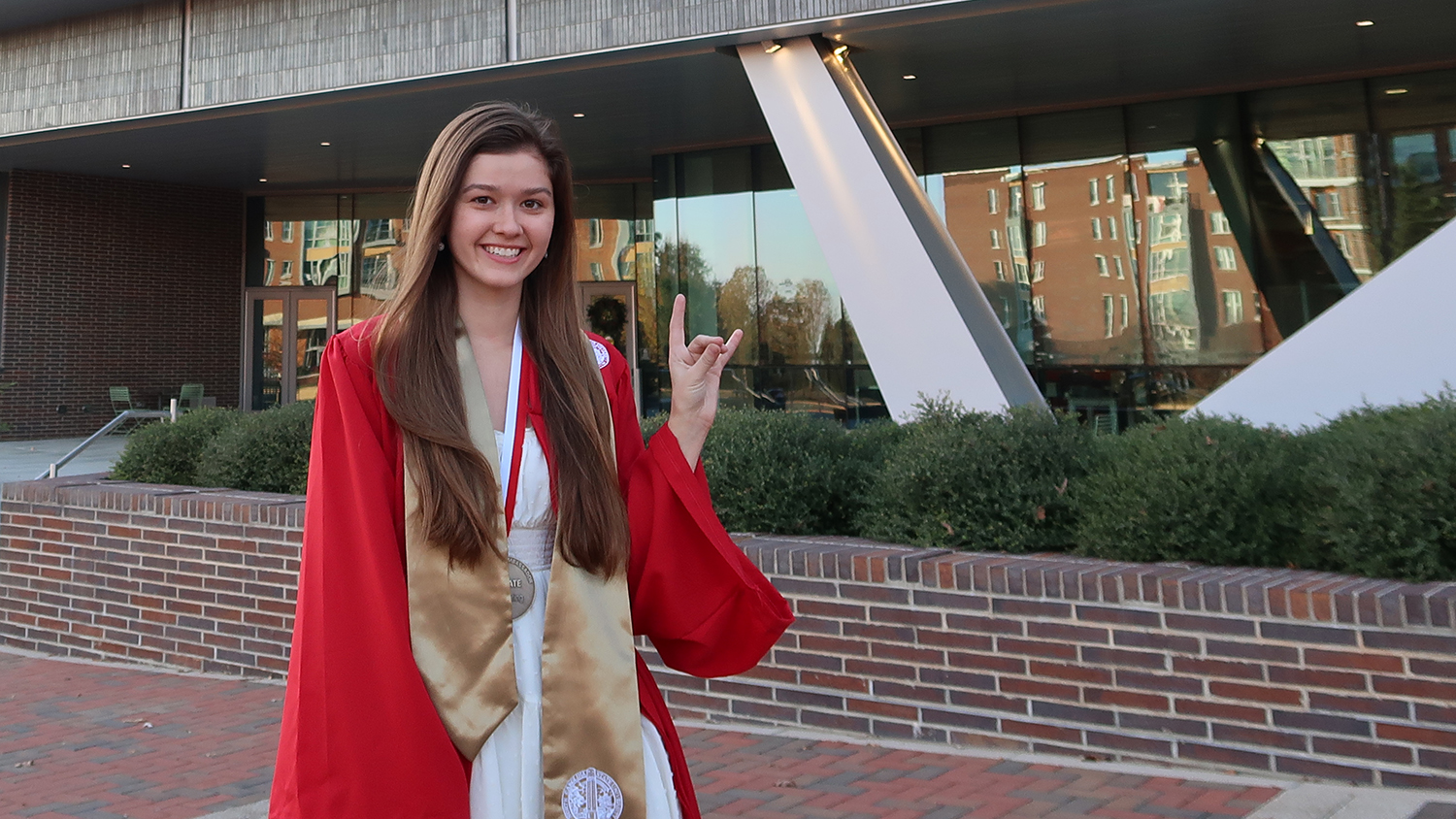 Sammi Easton, wearing a red graduation gown, flashes the NC State wolf sign in front of Fitts-Woolard Hall.
