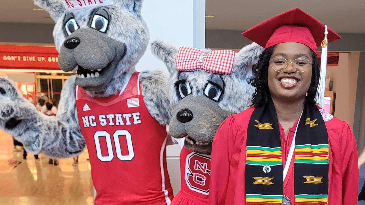 Maya Clinton smiles while wearing red graduation cap and gown. She is standing next to Mr. and Mrs. Wuf, NC State University mascots.