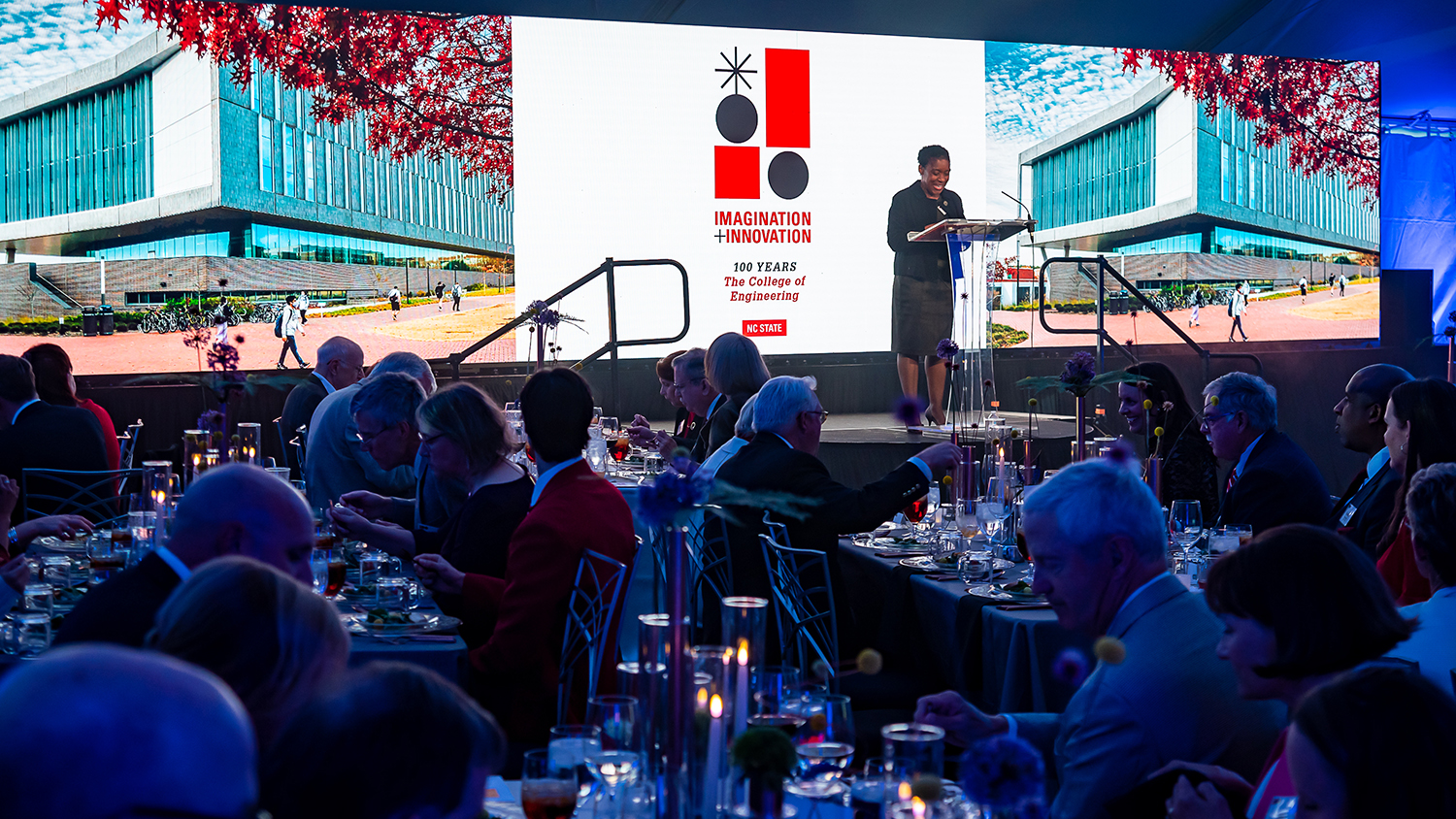 President Deborah B. Young, CE ‘77, speaks to attendees during the College of Engineering 100th anninversary celebration in May 2023. A large LED screen is behind her showing an image of Fitts-Woolard Hall and the 100th anniversary logo designed for the College of Engineering.