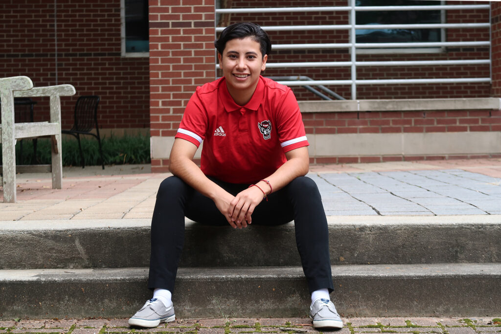 Daisy Aguilar Aguilar poses for a photo while sitting on the curb of an NCSU street. She is wearing a NC State red photo shirts and black pants.
